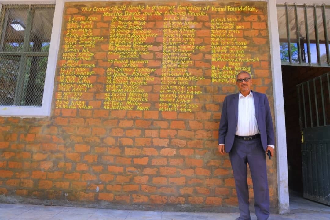 Erkab coordinator standing proudly before the Kamal Foundation donor recognition wall honoring community supporters